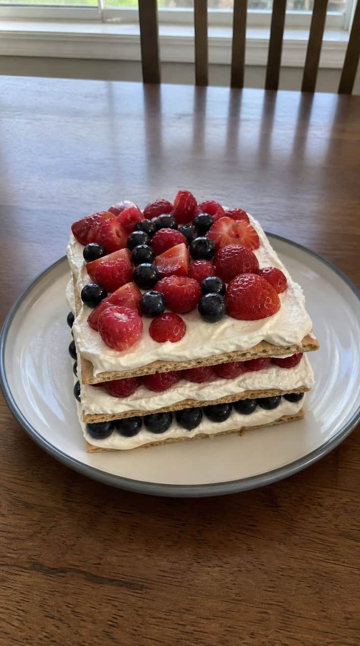 A layered icebox cake topped with whipped cream and fresh red and blue berries on a white plate.