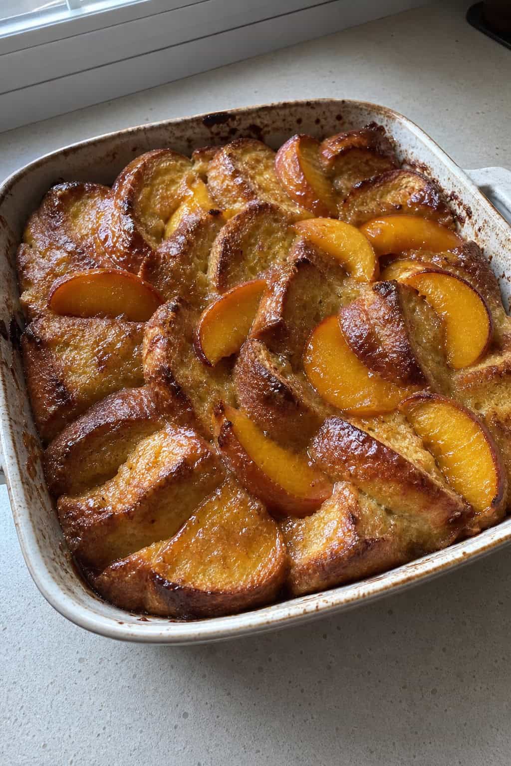 A freshly baked peach and bourbon French toast casserole in a rustic ceramic dish on a wooden table, with sliced fresh peaches and a sprig of herbs placed alongside for garnish.