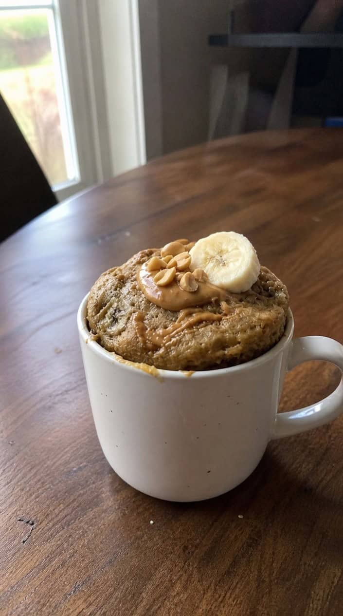 A ceramic mug filled with a dense, warmly golden peanut butter banana mug cake on a clean white surface, with a halved ripe banana and a spoonful of peanut butter and a few whole peanuts arranged beside it.