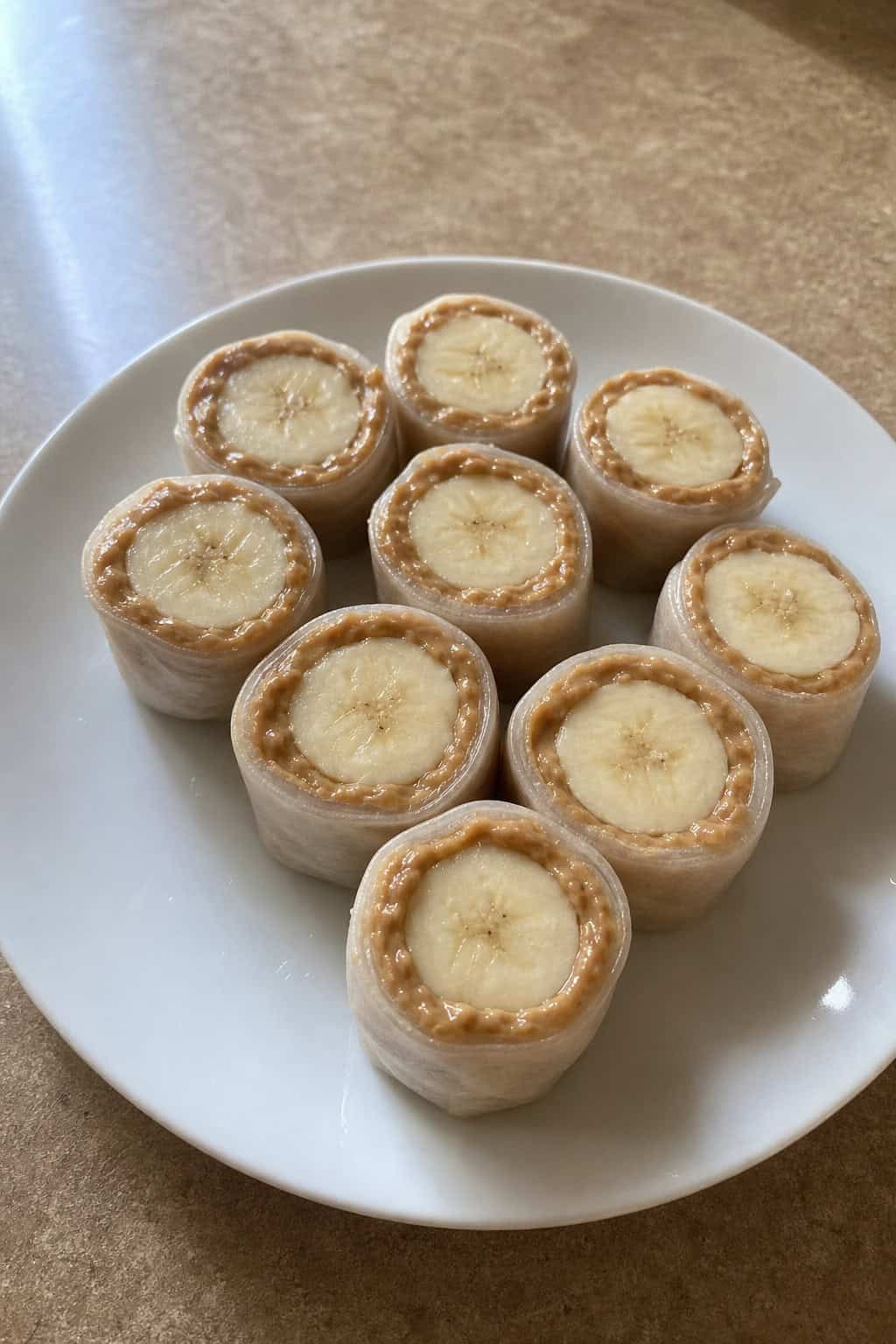 A plate of peanut butter banana sushi rolls arranged on a wooden table with a bowl of peanut butter and a banana in the background.