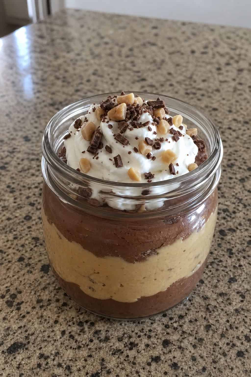A glass jar filled with peanut butter chocolate cheesecake pudding topped with whipped cream and chocolate shavings on a wooden surface.