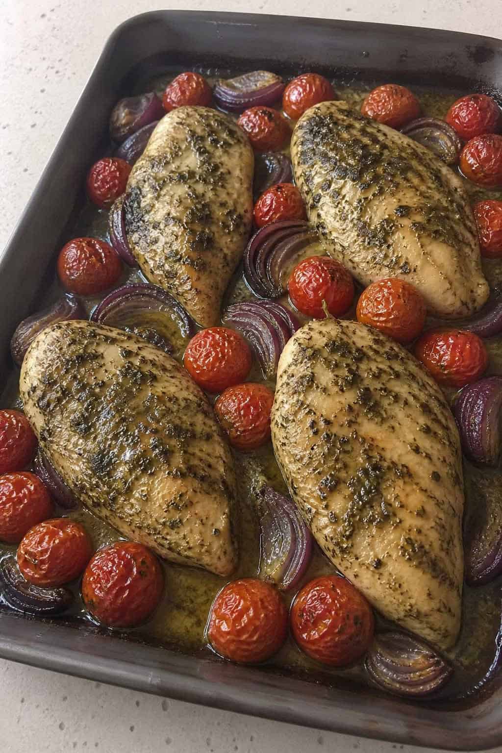 Pesto-slicked chicken breasts with blistered cherry tomatoes on a foil-lined sheet pan sitting on a light kitchen countertop.