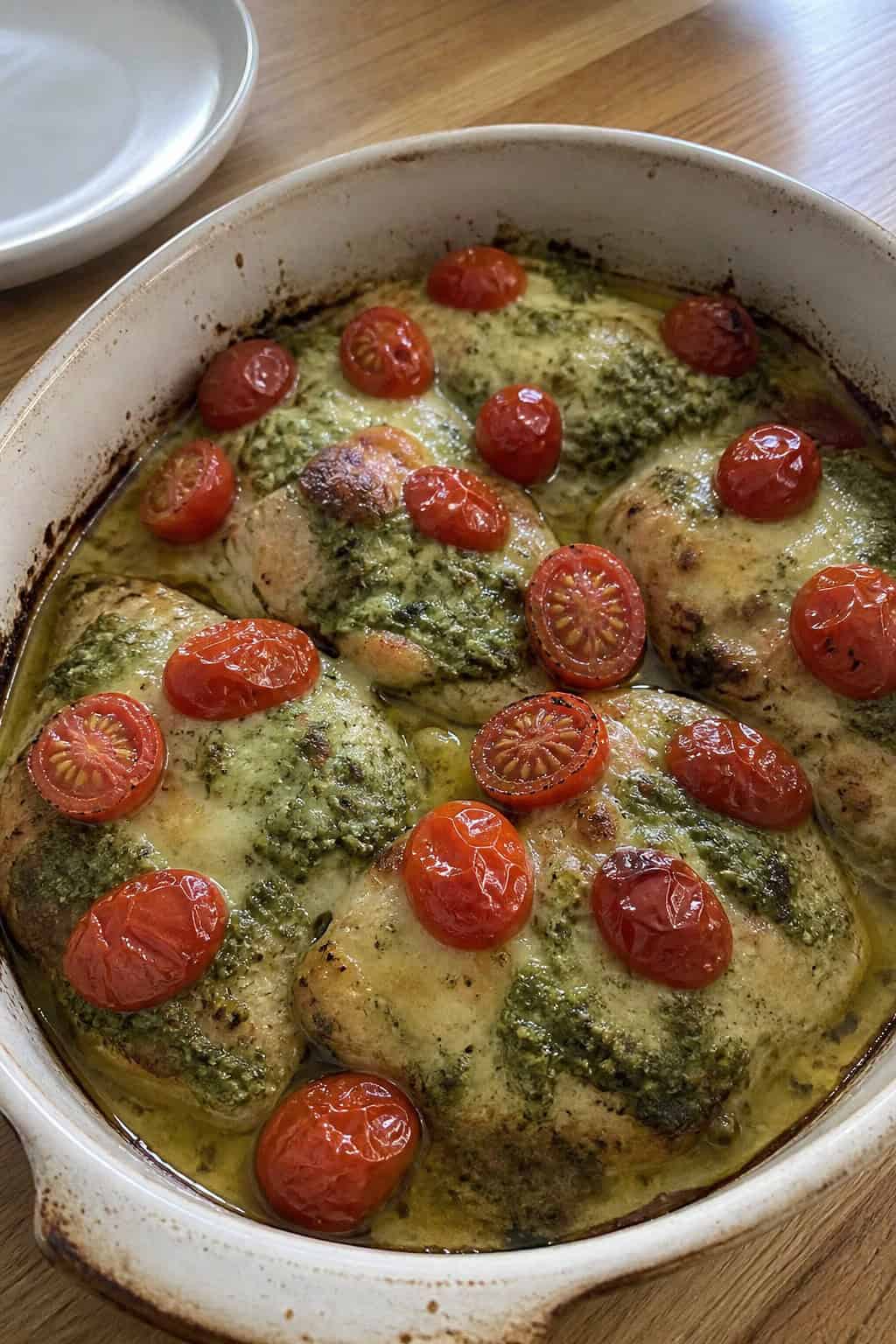 20 Chicken Casserole Recipes for Big Gatherings 12 A baked chicken and cherry tomato casserole in a ceramic dish on a wooden table, with golden mozzarella and Parmesan topping and fresh basil leaves visible.