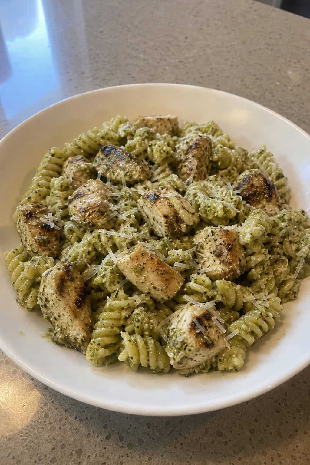 A bowl of rotini pasta with pesto sauce and grilled chicken garnished with basil and Parmesan cheese on a wooden table.