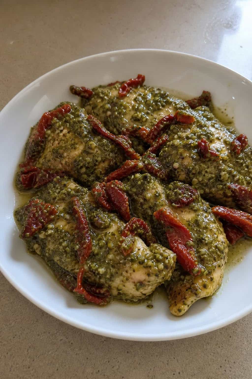 A bowl of pesto chicken topped with sun-dried tomatoes and fresh basil on a wooden table.