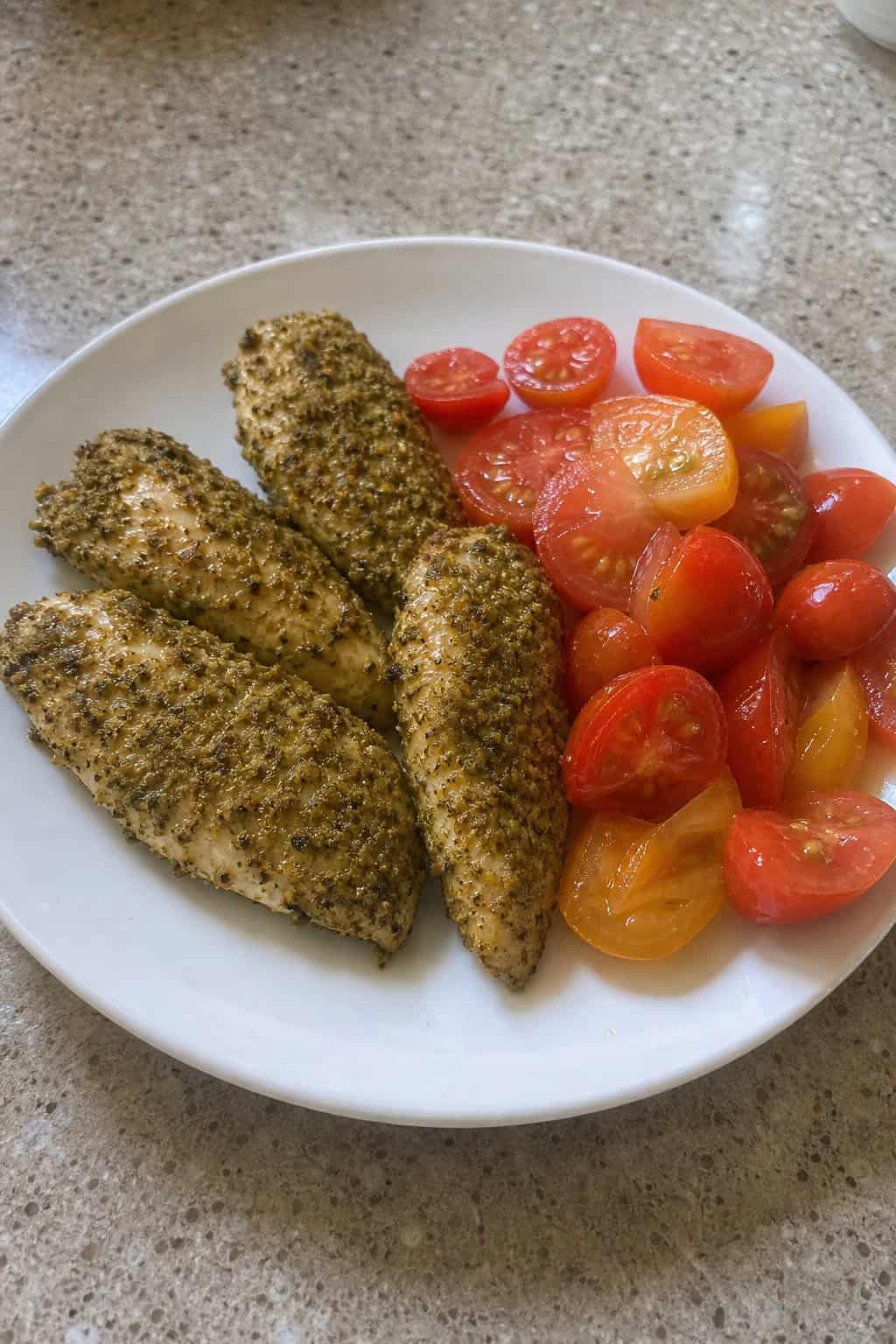 Plate of pesto-crusted chicken tenders served with a fresh tomato salad on a wooden table.