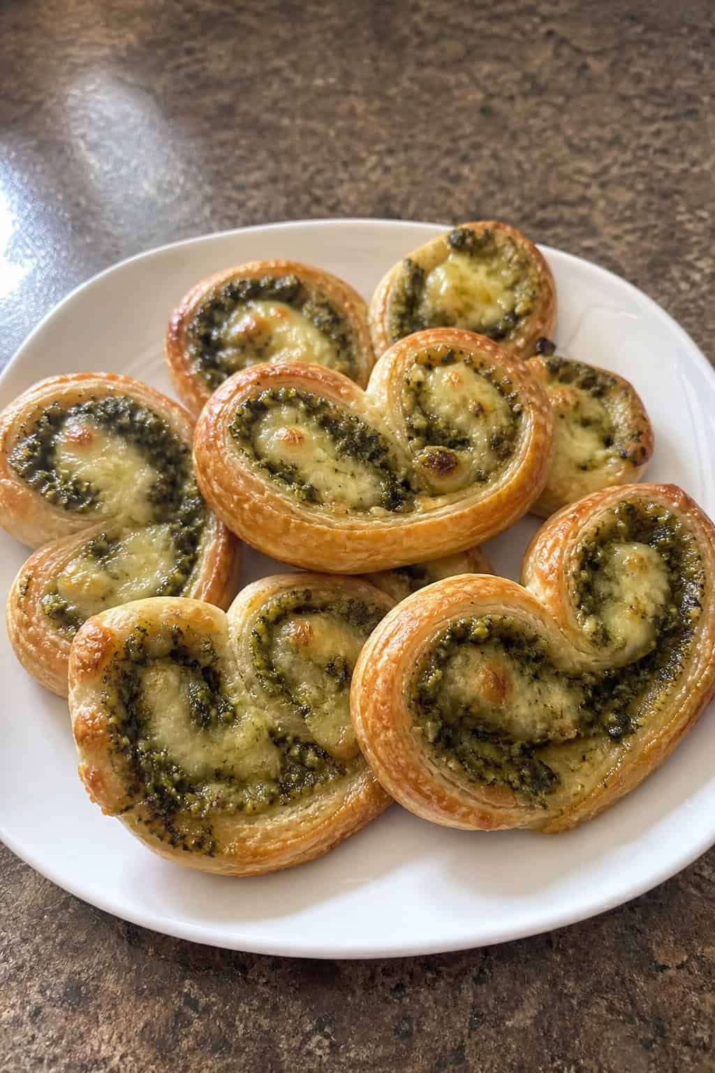 A plate of golden brown pesto-mozzarella palmiers garnished with basil leaves on a wooden table with a bowl of pesto and a glass of white wine in the background.