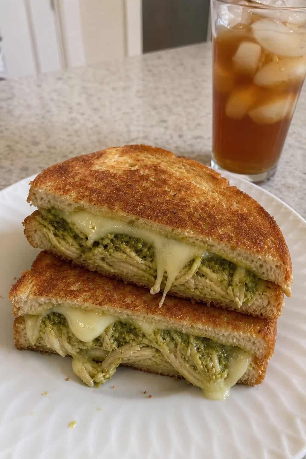 A grilled cheese sandwich on a wooden board, cut diagonally to show layers of melted mozzarella and pesto-coated shredded chicken between golden-brown sourdough slices, with fresh basil leaves and a glass of iced tea in the background.