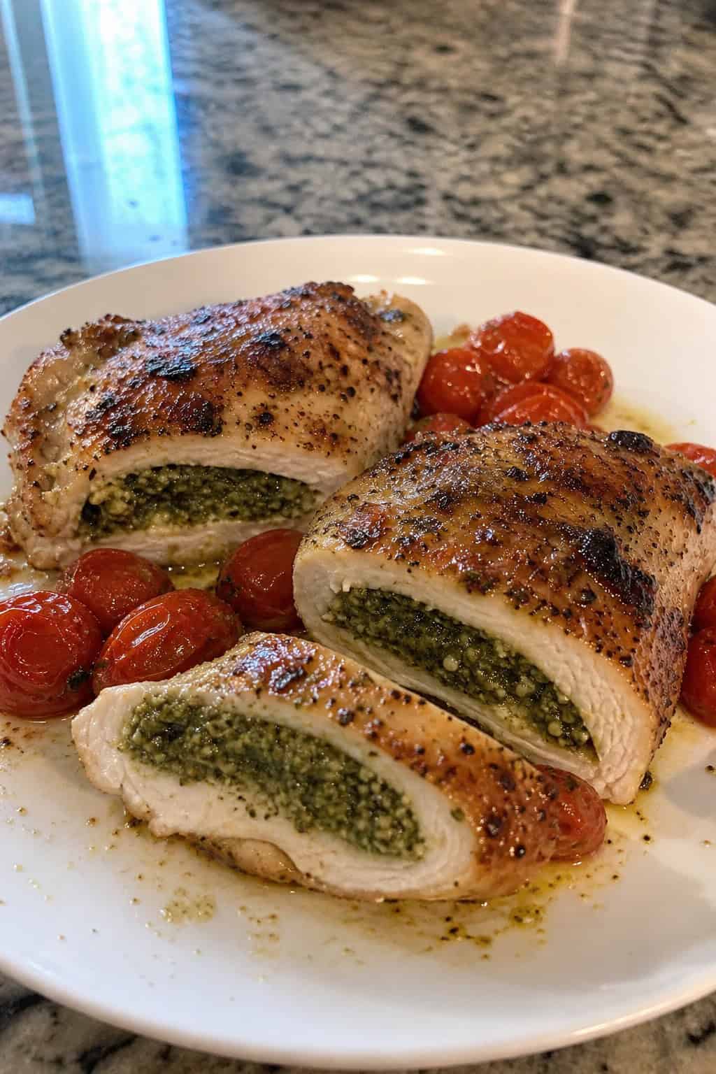 A plate of golden brown chicken thighs stuffed with green pesto, garnished with basil leaves and roasted cherry tomatoes on a white plate.