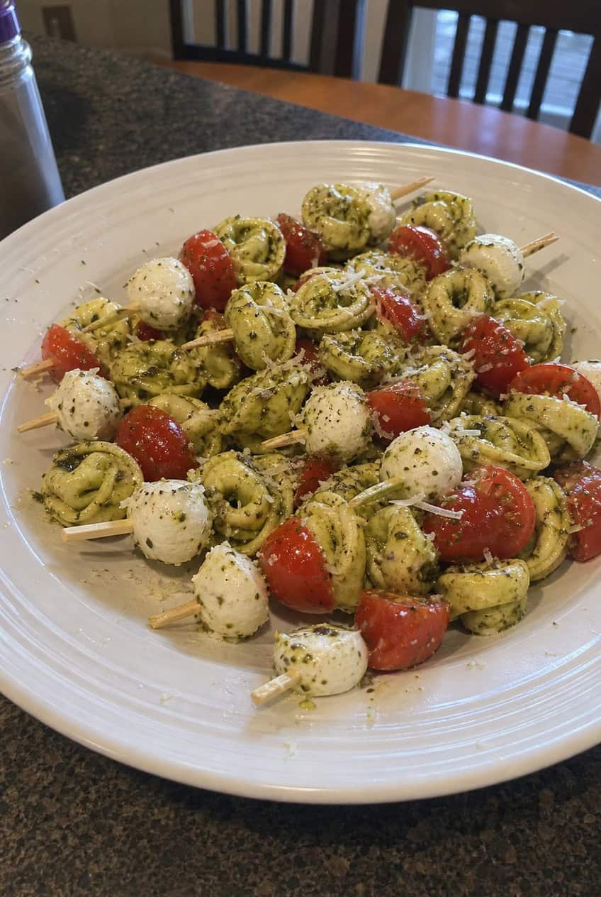 A close-up of wooden skewers on a board, each holding a plump cheese tortellini, a halved red cherry tomato, and a small mozzarella ball coated in glossy green pesto.