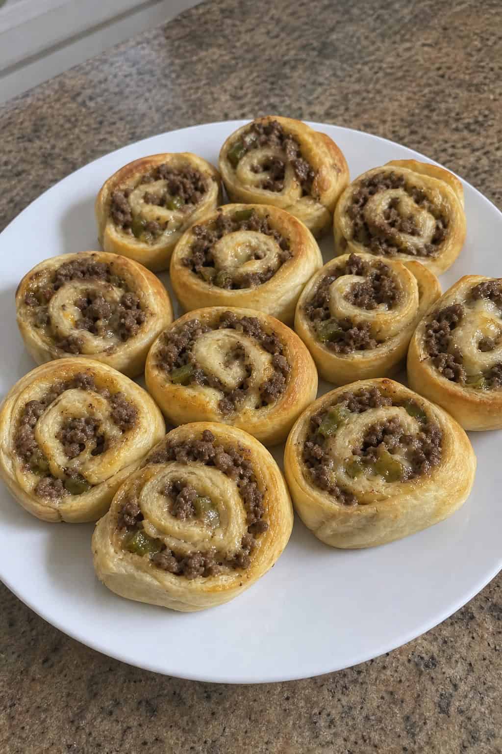 A wooden board with several Philly cheesesteak pinwheels showing thinly sliced beef, melted provolone, and caramelized onions inside flour tortilla rolls, with small dipping sauce bowls and game day items in the background.