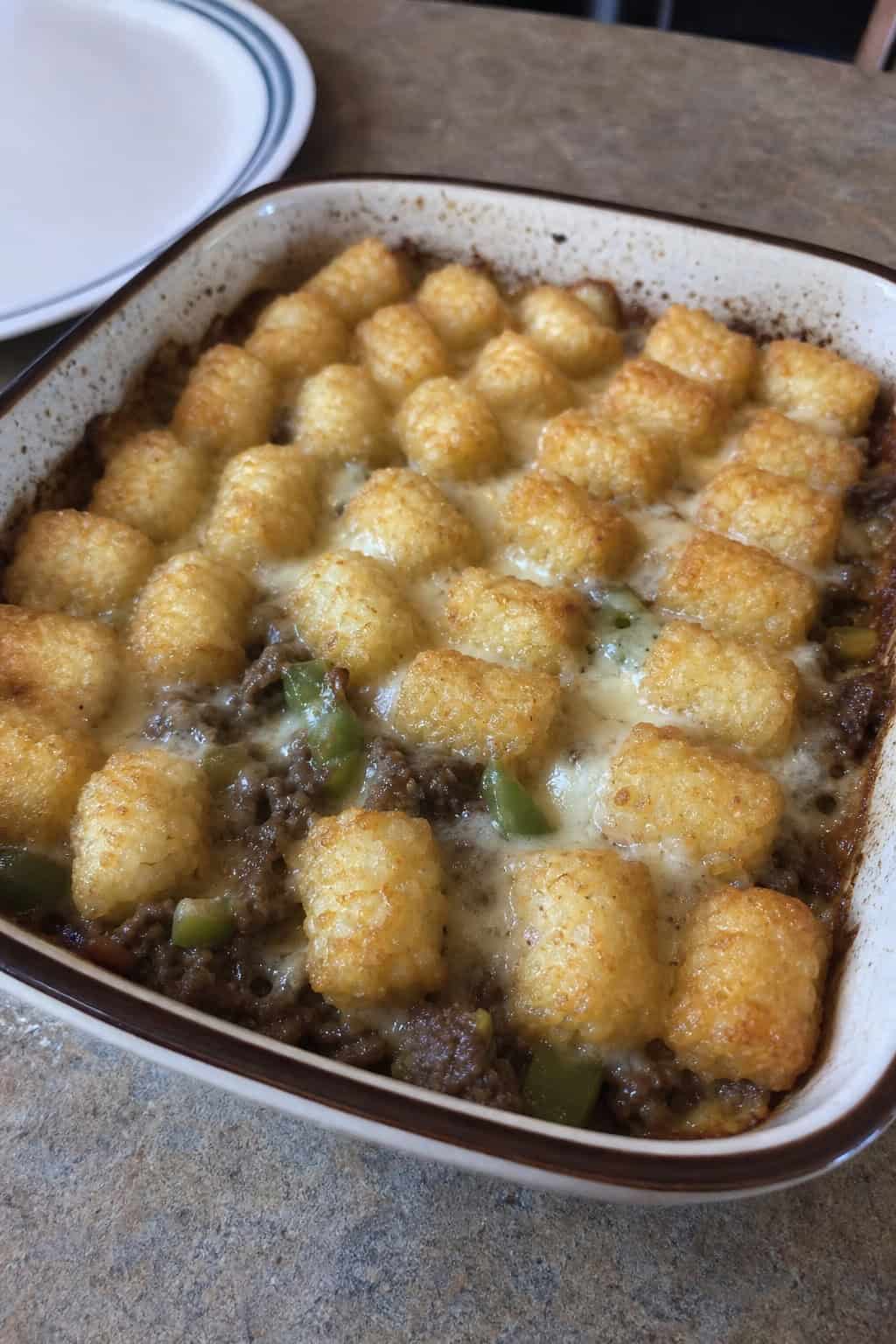 A baked Philly cheesesteak tater tot casserole in a ceramic dish with golden tots on top and green bell pepper and onion visible where the filling meets the edges, set on a wooden table.
