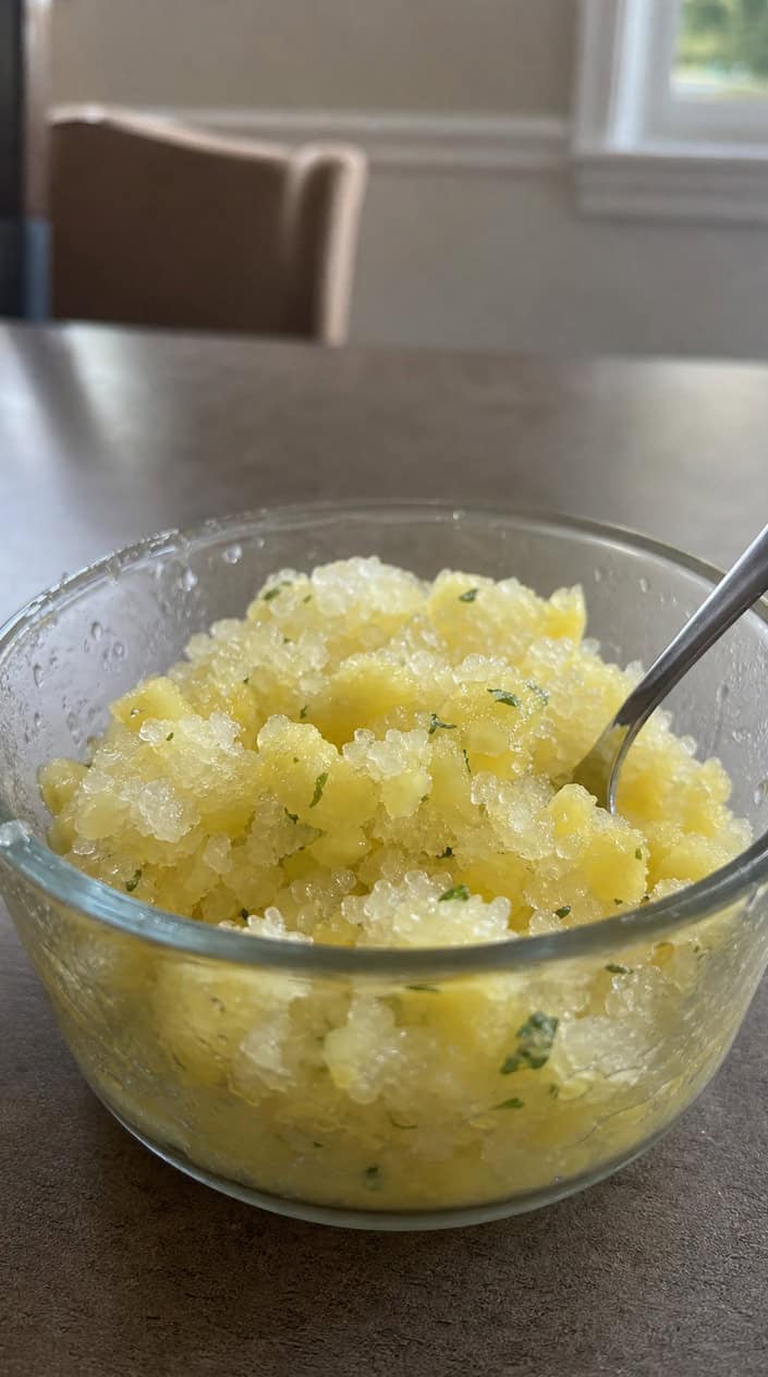 A bowl of pineapple and mint granita garnished with fresh mint leaves on a wooden table in a sunlit backyard.