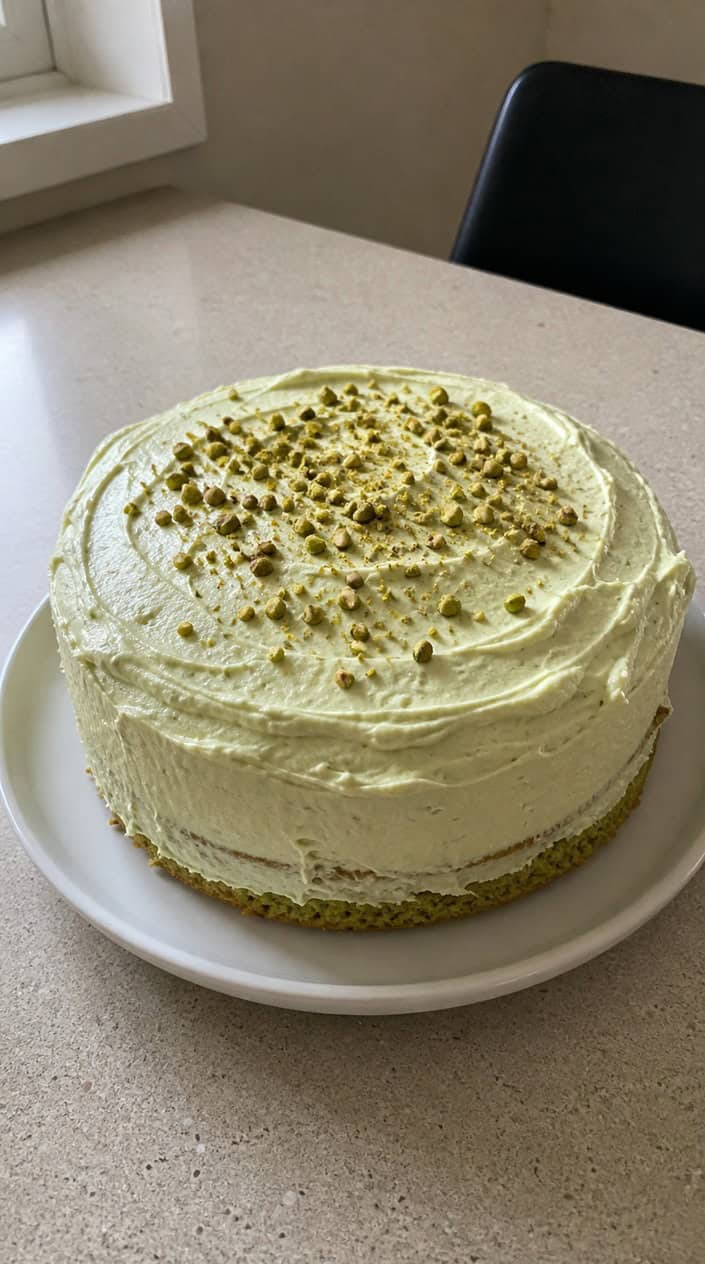 A pistachio sponge cake with pale green whipped frosting on a white plate on a wooden surface.
