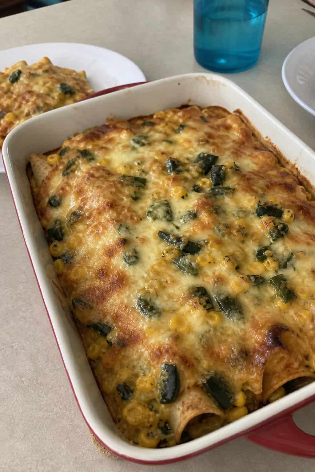 A baked poblano and corn enchilada casserole with a golden bubbling queso-style cheese topping in a ceramic dish, with halved fresh poblano peppers and corn kernels arranged on the wooden surface around it.