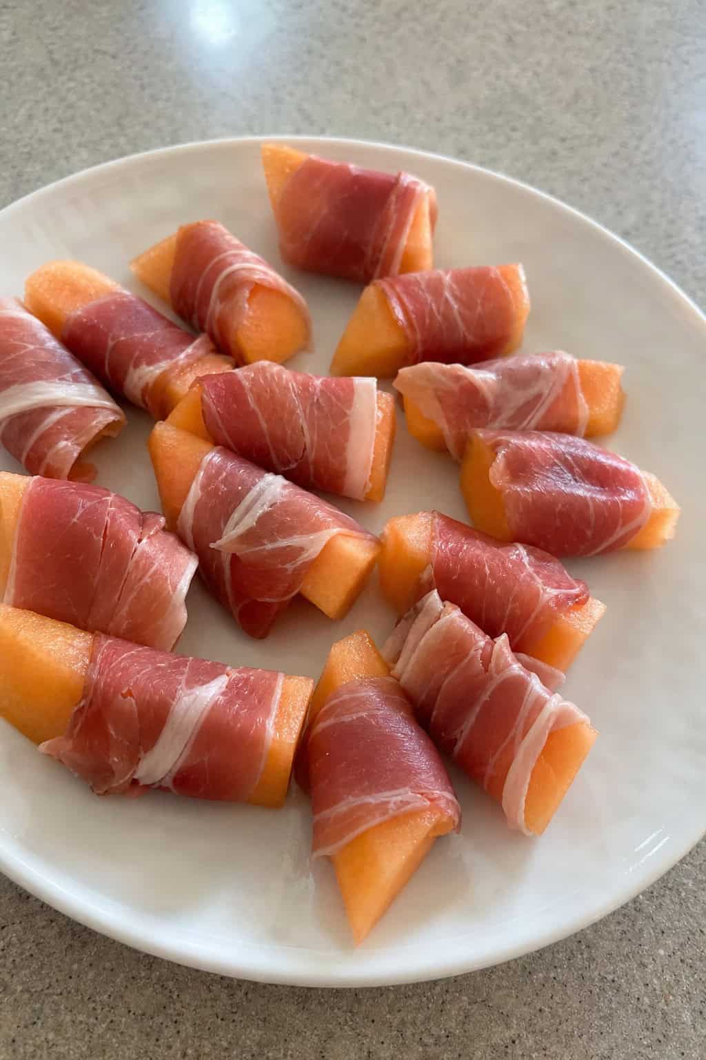 Close-up of prosciutto-wrapped melon bundles on a white platter with mint garnish on a wooden table.