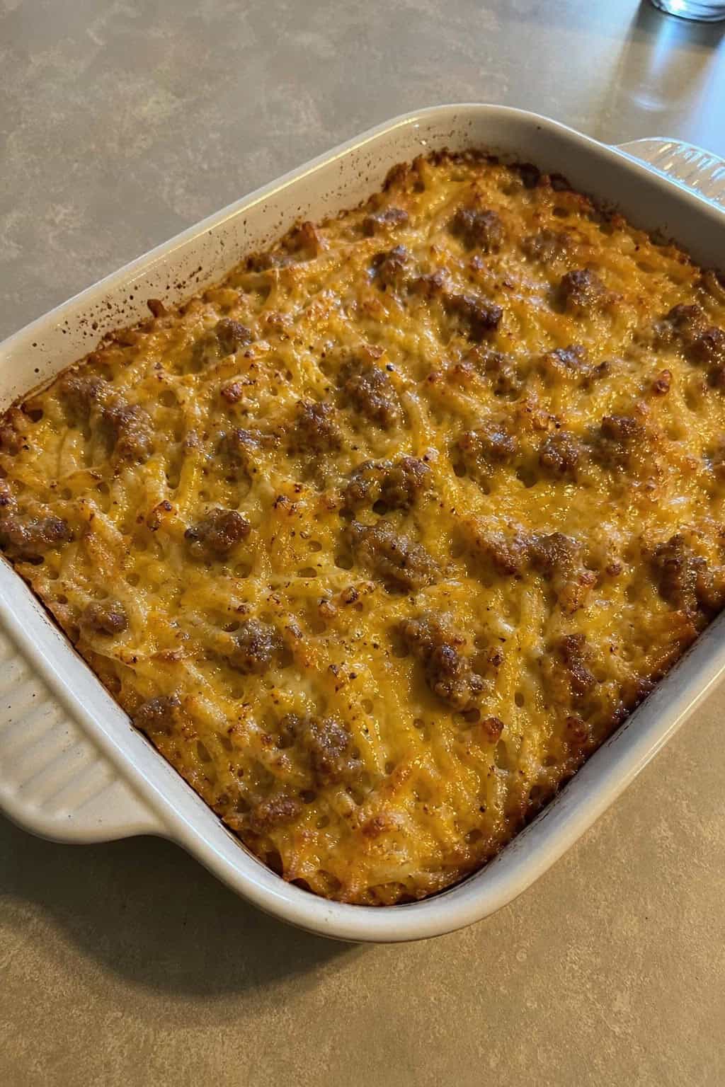 A golden-brown turkey sausage and hashbrown casserole in a white ceramic dish on a wood table with a glass of orange juice and a small bowl of fresh berries to the side.
