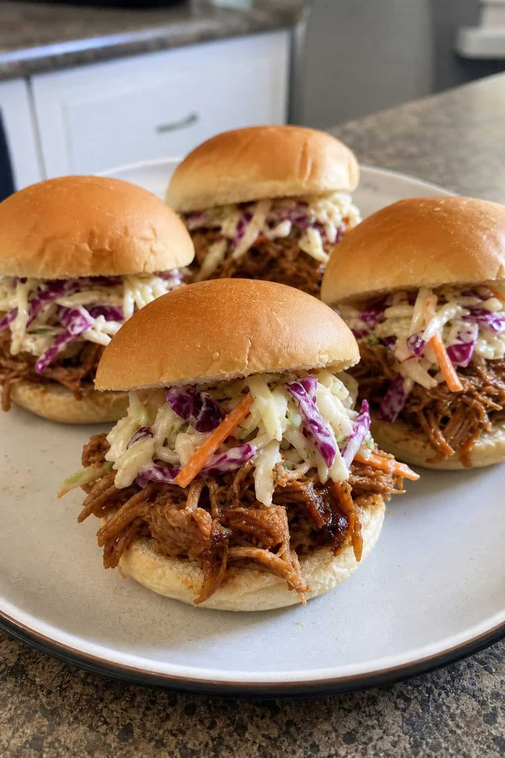 Close-up of pulled pork sliders topped with coleslaw on a wooden board with a bowl of coleslaw beside them.