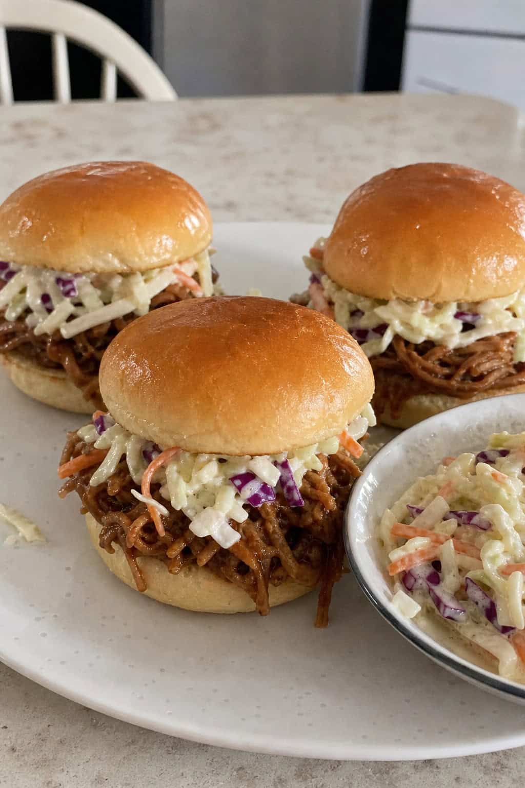 A plate of pulled pork sliders topped with coleslaw on a wooden board in a kitchen setting.