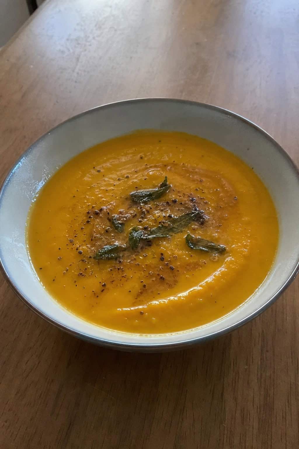 A bowl of pumpkin, sage, and brown butter soup garnished with sage leaves on a wooden table with pumpkins and herbs around it.