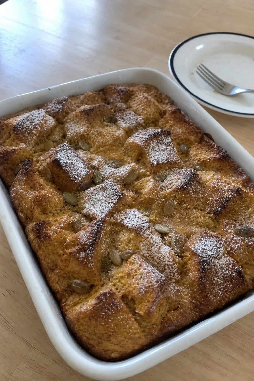A pumpkin-spice breakfast casserole in a deep ceramic dish on a wooden table with small decorative pumpkins, cinnamon sticks, and rust-colored autumn leaves in the background.