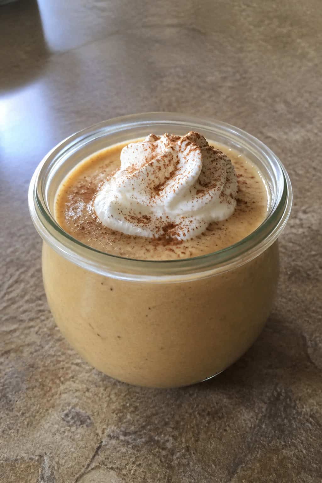 A glass jar filled with pumpkin spice cheesecake pudding topped with whipped cream and cinnamon, surrounded by autumn decorations on a wooden table.