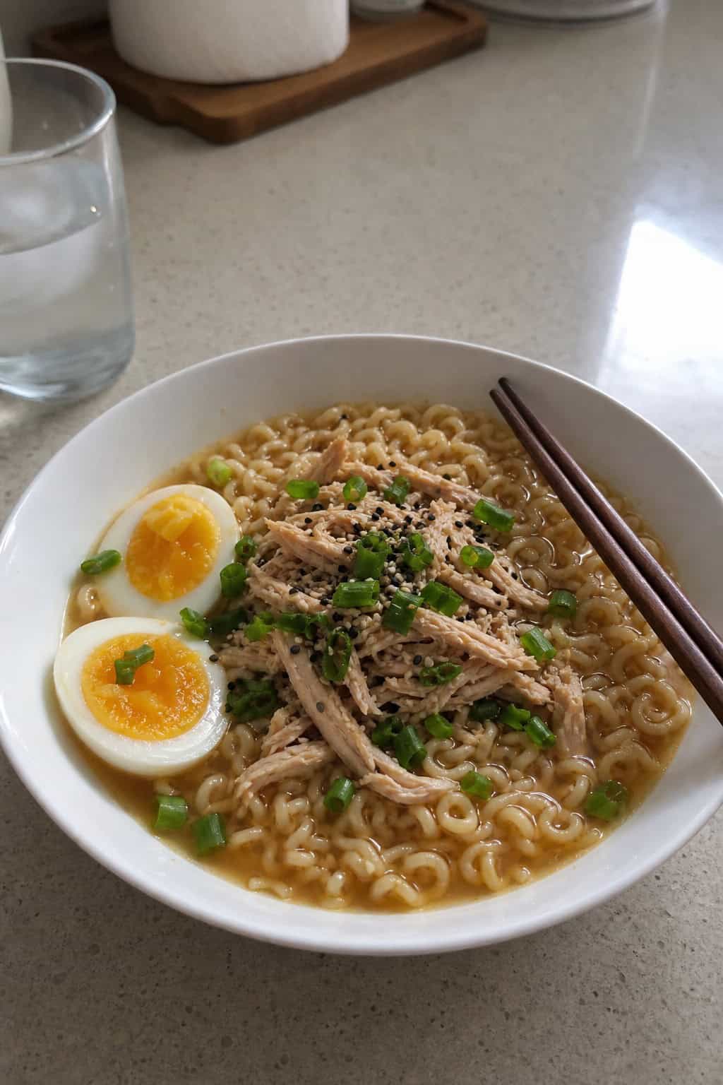 A bowl of ramen noodles topped with shredded chicken, boiled eggs, and green onions on a wooden countertop with chopsticks.