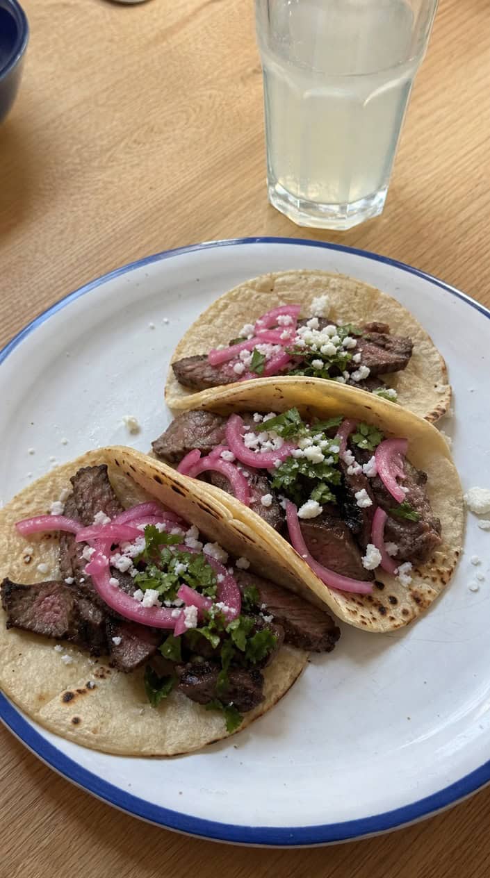 A plate of flank steak tacos topped with pickled onions, garnished with cilantro and lime wedges on a wooden platter.