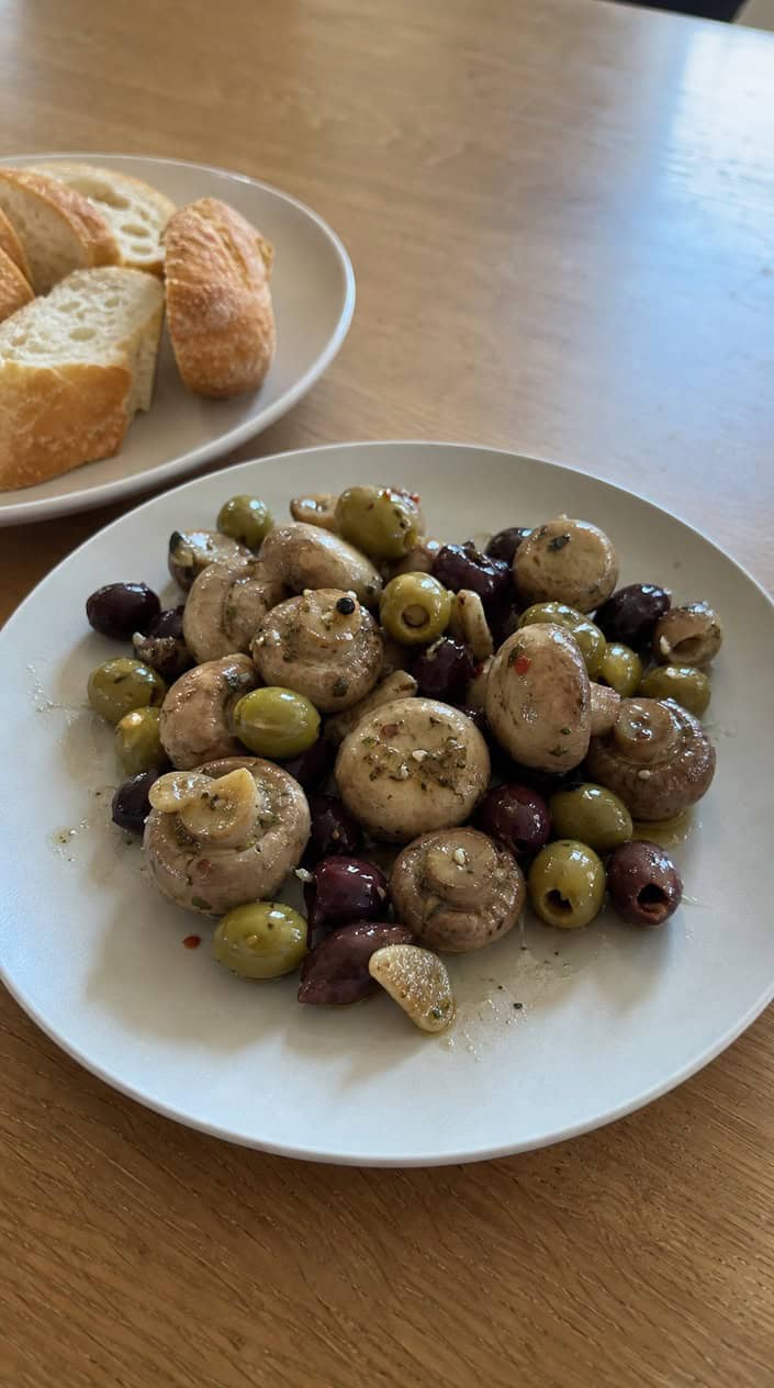 A wooden board with marinated mushrooms and olives, garnished with fresh herbs and accompanied by sliced bread and small bowls of olive oil.