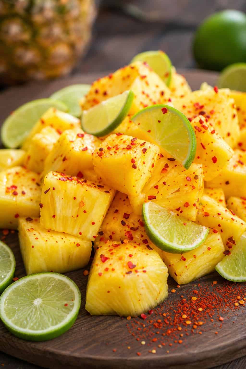 Close-up of pineapple chunks sprinkled with chili flakes and served with lime wedges on a wooden board.