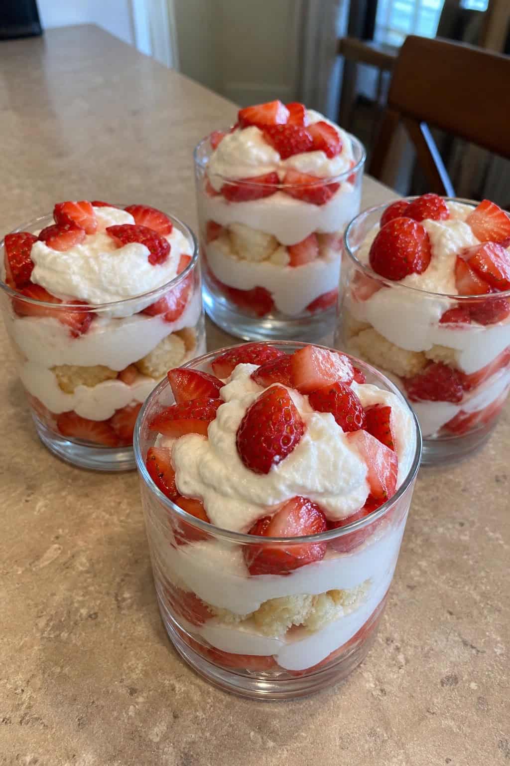 Three glass cups filled with layers of sliced strawberries, whipped cream, and shortcake pieces on a wooden table.