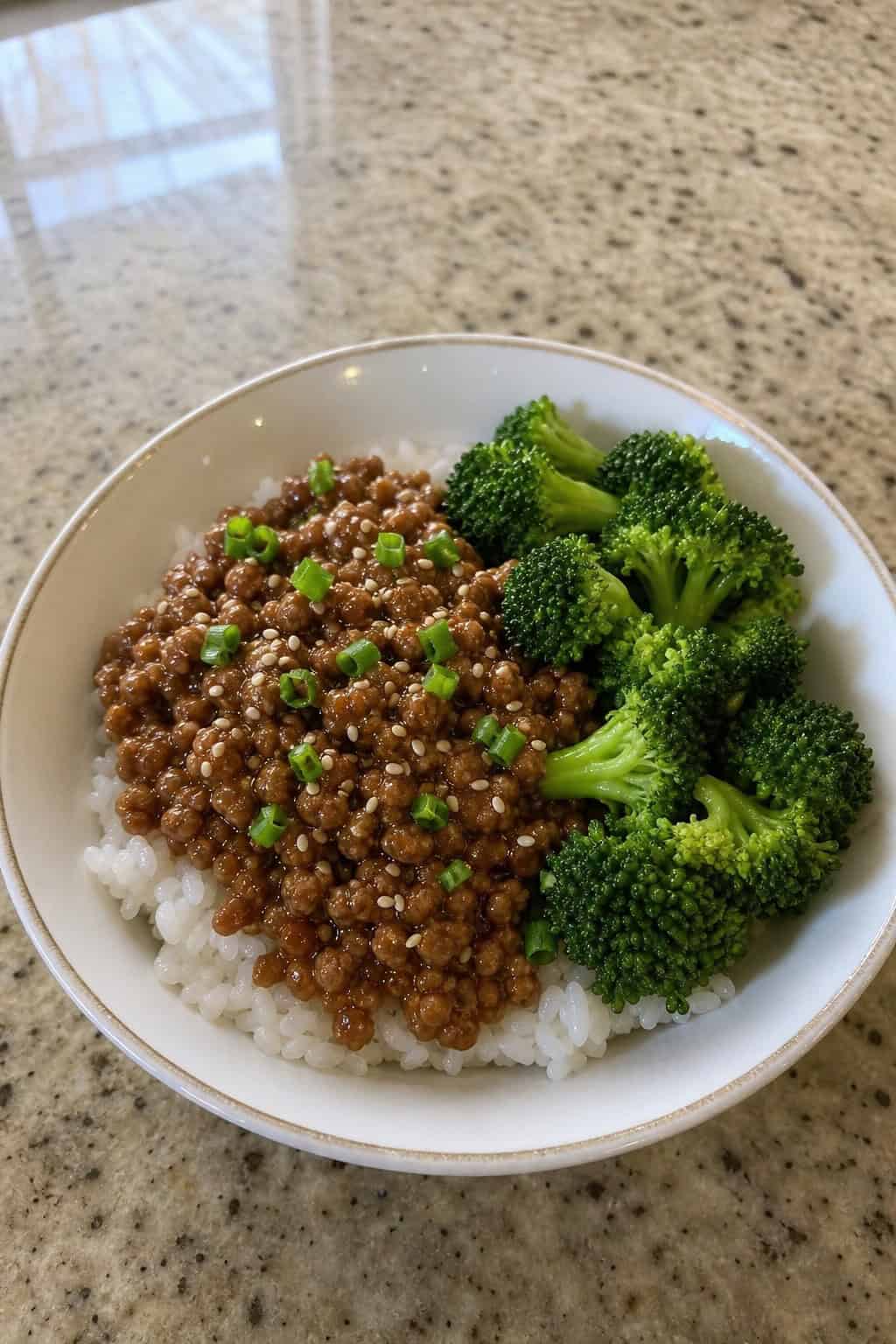 A bowl of minced chicken teriyaki with steamed broccoli and white rice on a wooden table.