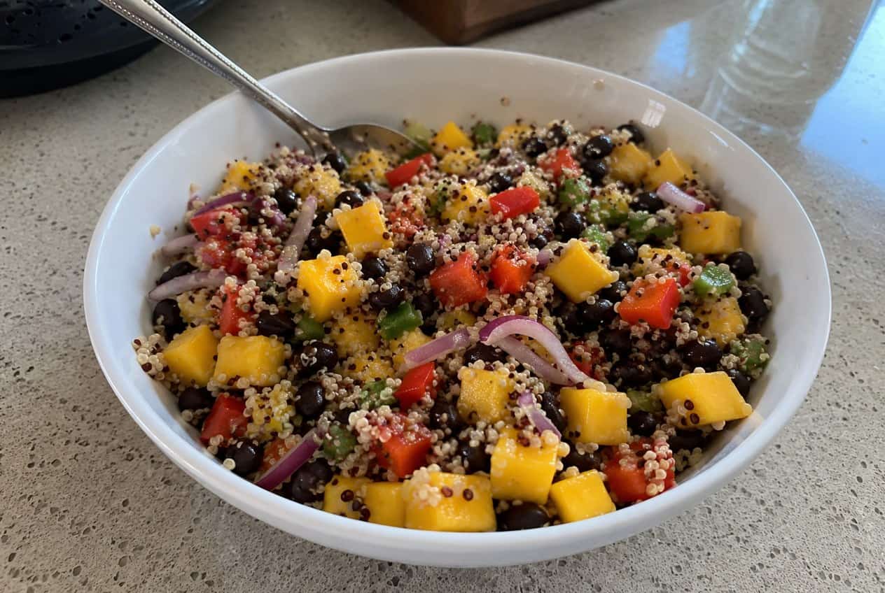 A deep bowl packed with fluffy white quinoa, bright orange mango cubes, black beans, diced red bell pepper, chopped red onion, and fresh cilantro on a wooden table.