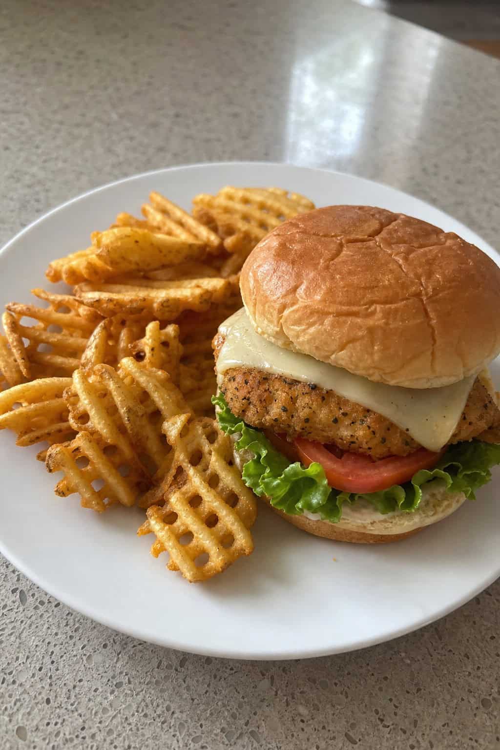 A ranch-seasoned chicken tender sandwich with shredded lettuce, cheese, and tomato on a toasted bun, served beside a pile of crispy waffle fries on a plate