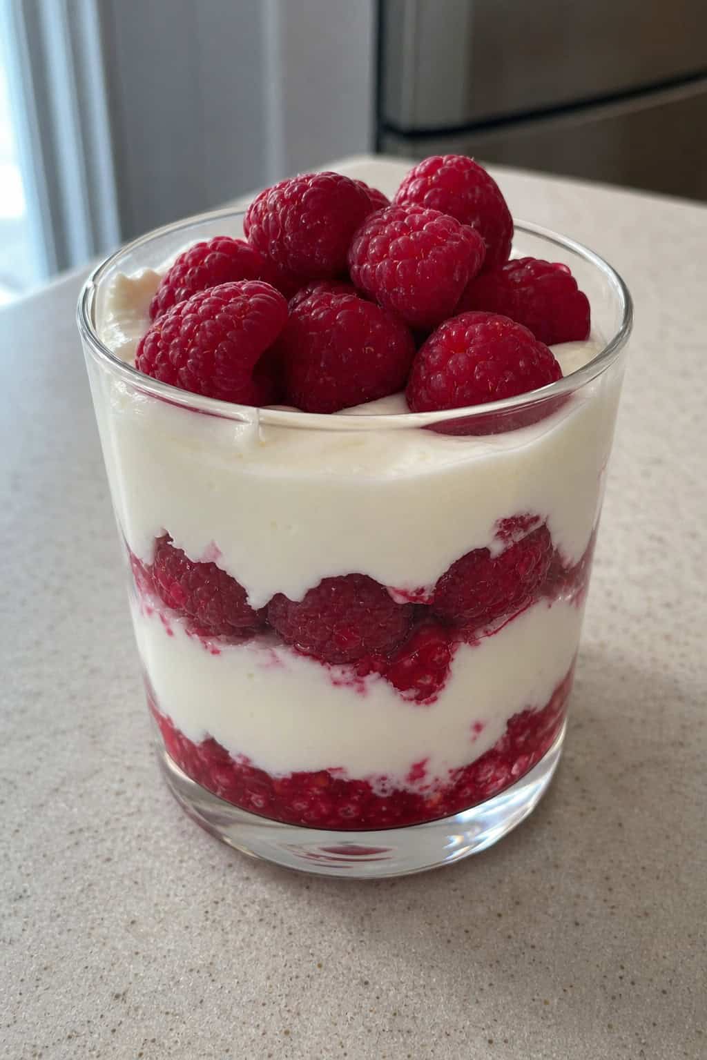A glass cup filled with raspberry white chocolate keto cheesecake pudding topped with fresh raspberries and a mint leaf on a wooden table.