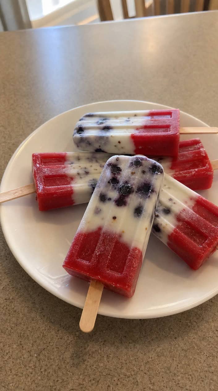 Red, white, and blueberry parfait popsicles arranged on a wooden table with fresh berries around them.