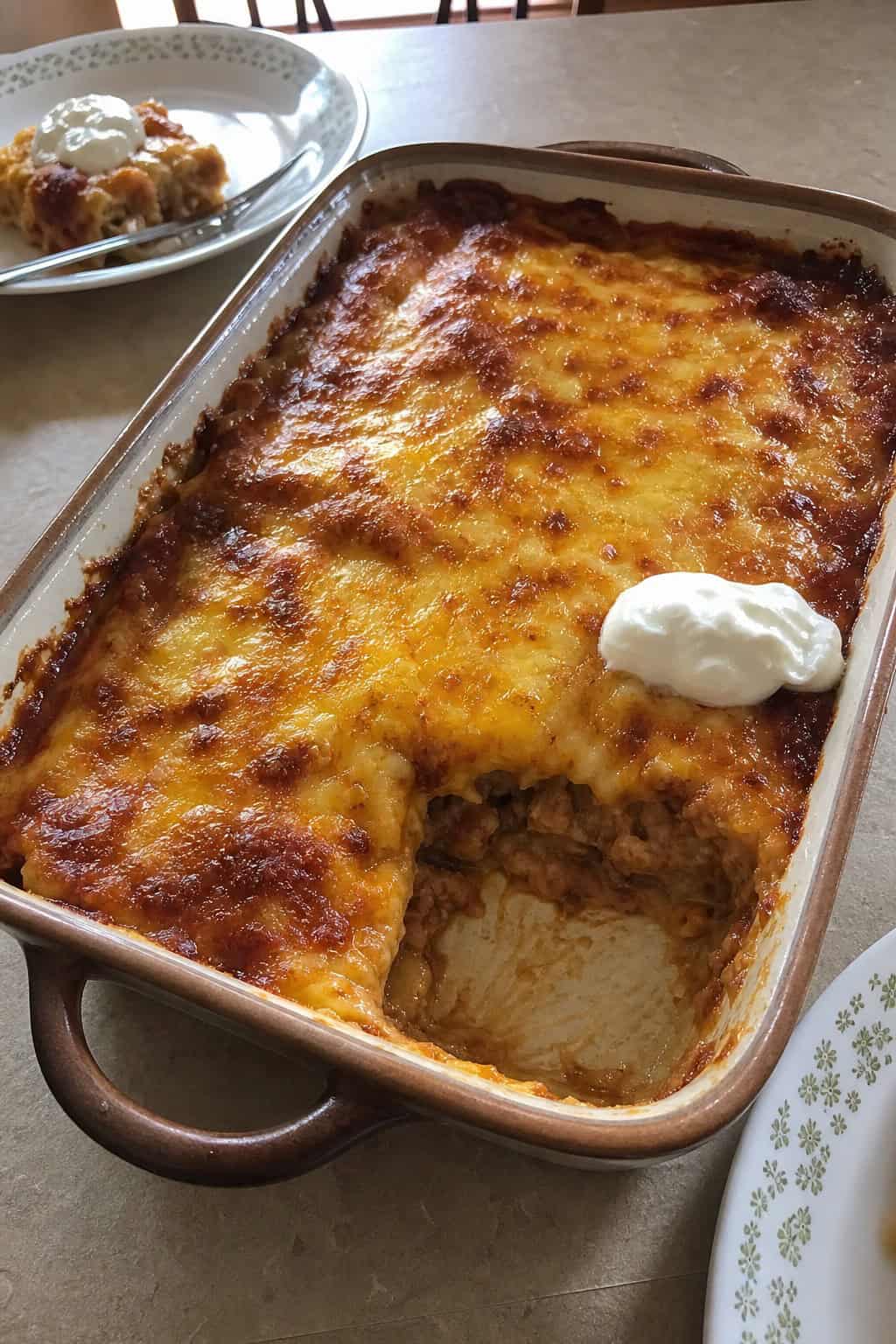 A baked refried bean and cheese enchilada casserole in a white ceramic dish with a deep golden cheese top and visible red sauce around the edges, served with a small bowl of sour cream on the kitchen table.