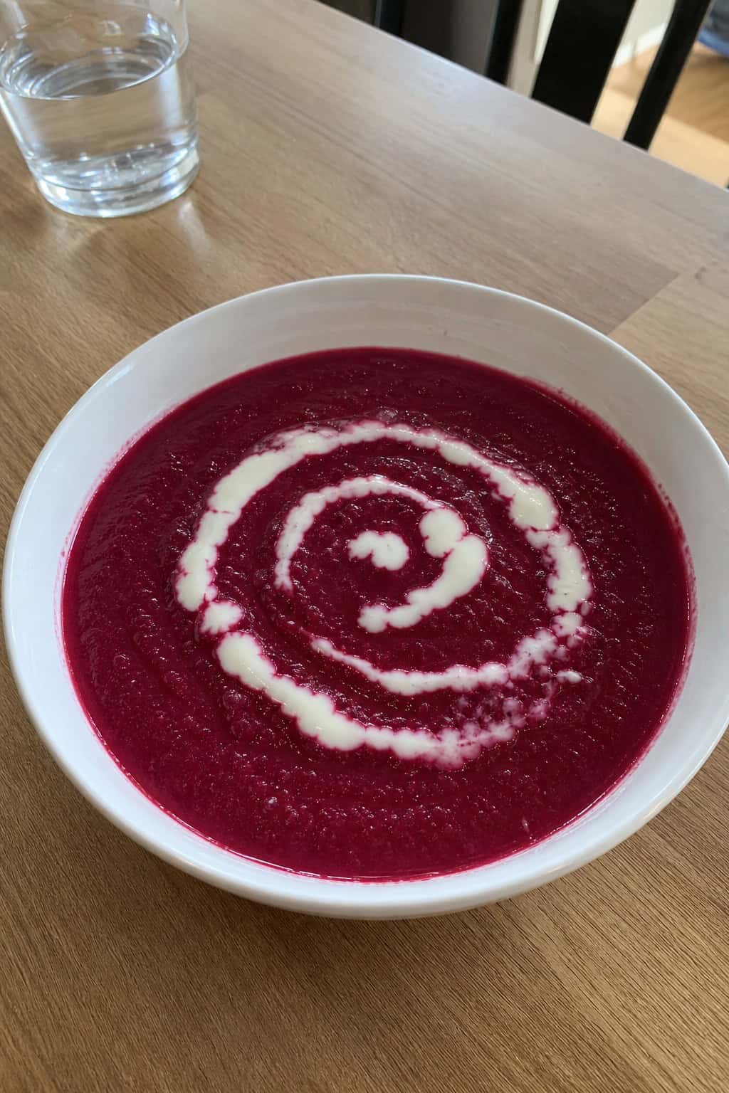 A bowl of roasted beet and apple soup garnished with horseradish sauce and fresh herbs on a wooden table, surrounded by fresh beets, apples, and horseradish.
