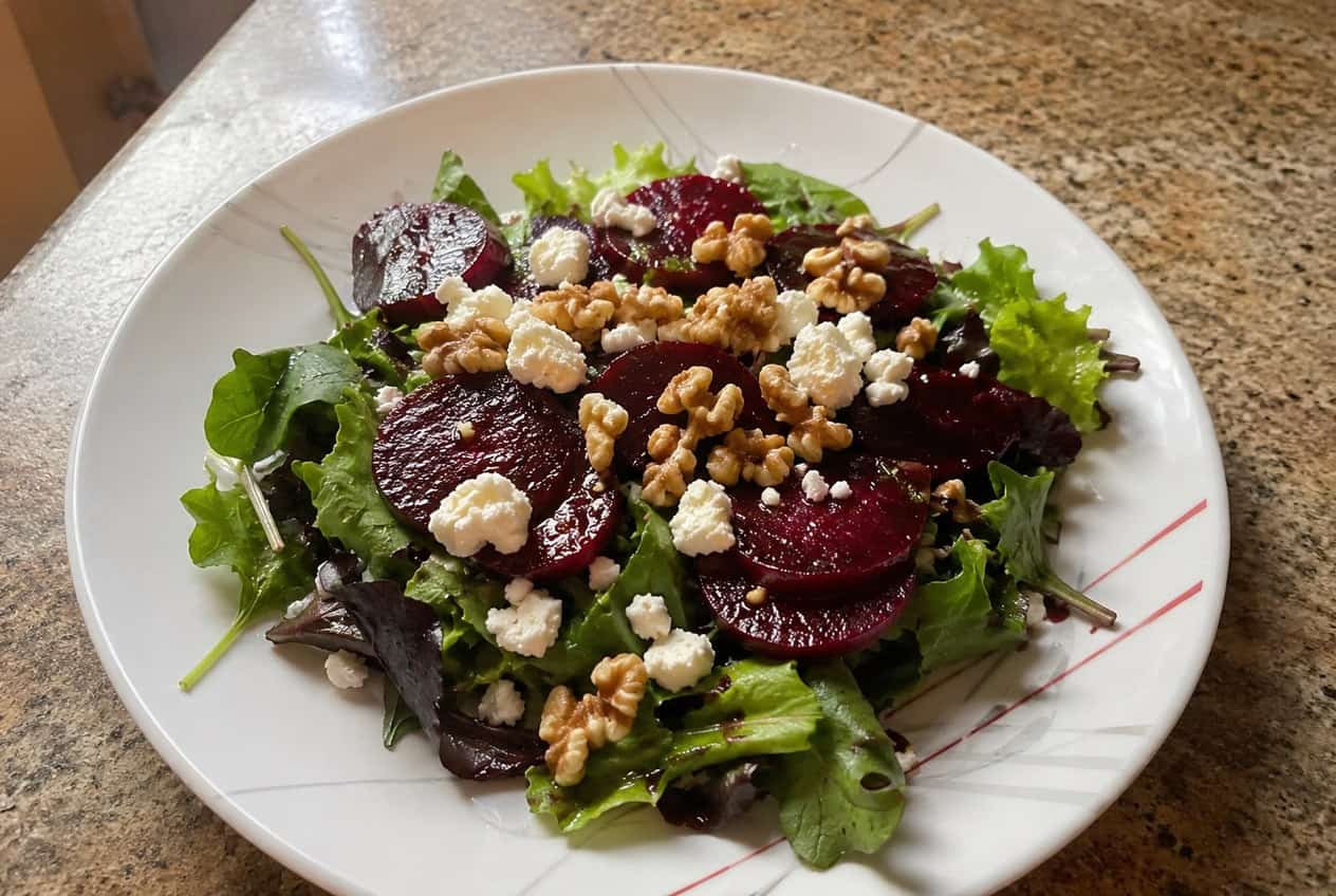 A wide plate of mixed dark greens topped with deep magenta roasted beet wedges, crumbled white goat cheese, and roughly chopped toasted walnuts, with a honey balsamic dressing pooling around the edges.