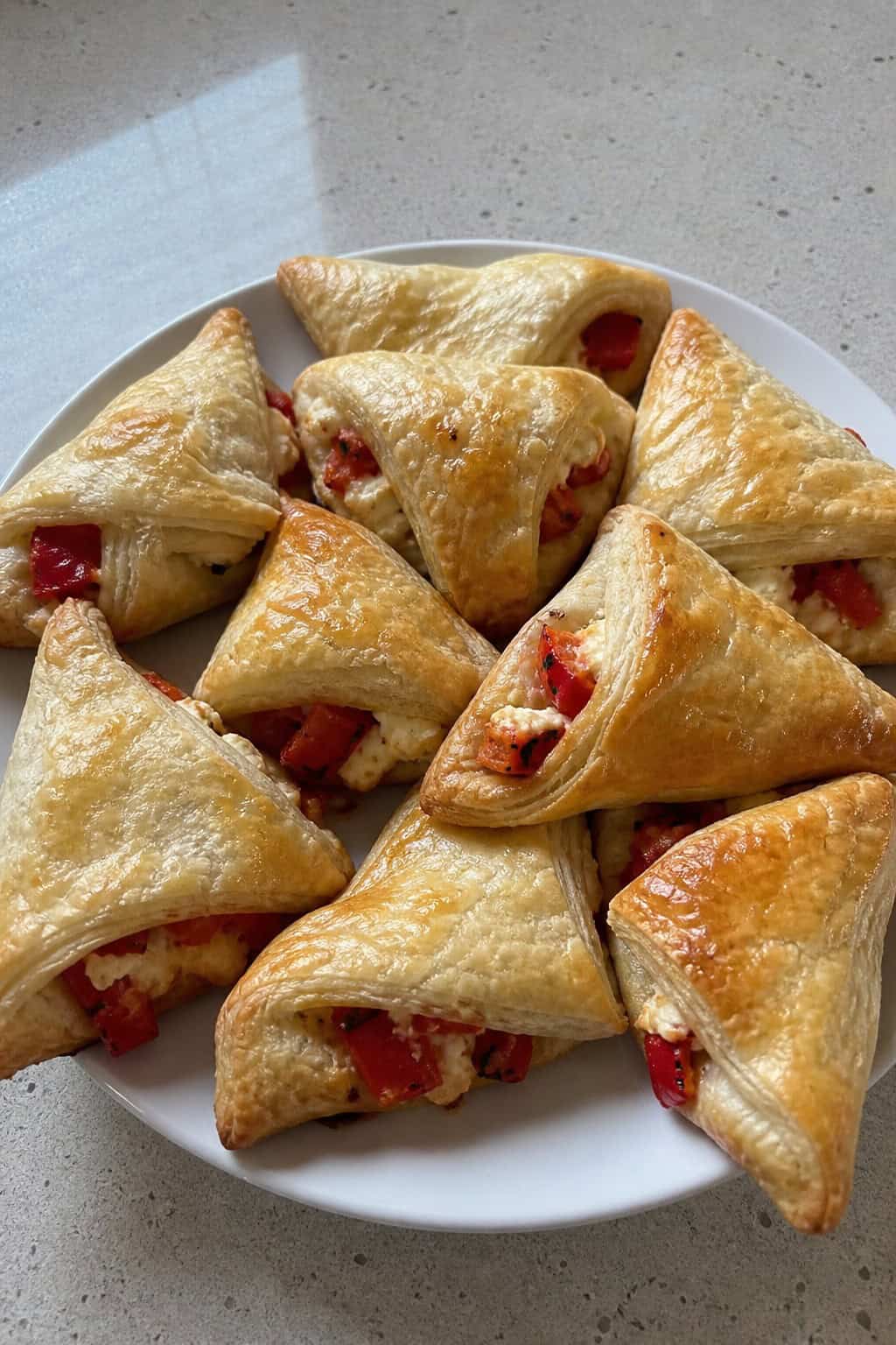 A wooden board with golden puff pastry triangles filled with roasted red peppers and feta, garnished with fresh herbs and a small bowl of dipping sauce.