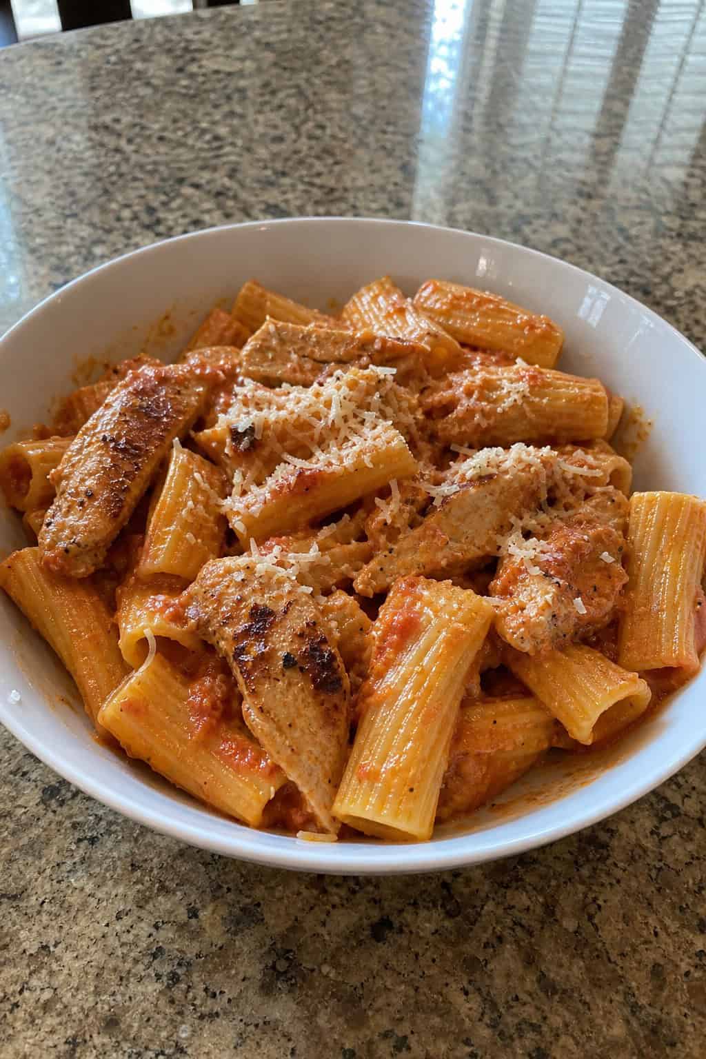 A bowl of roasted red pepper chicken rigatoni pasta garnished with basil and Parmesan cheese on a wooden table.
