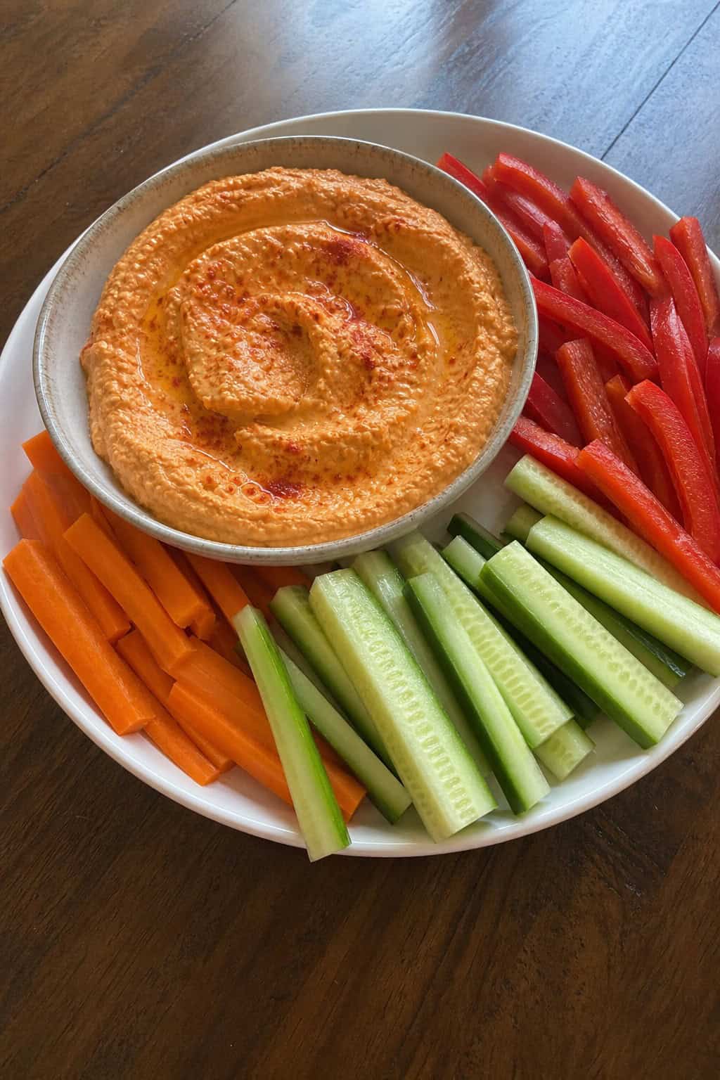 A bowl of roasted red pepper hummus surrounded by fresh vegetable sticks on a wooden table.