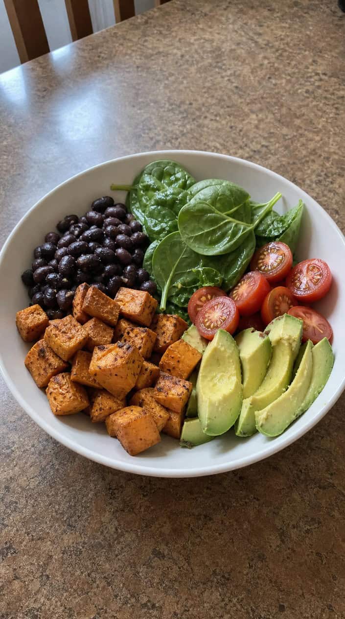 A wide bowl with a base of brown rice topped with caramelized cubed sweet potato, matte black beans, sliced avocado fanned across one side, a scattering of cherry tomatoes, and fresh cilantro, shot on a warm wood surface.