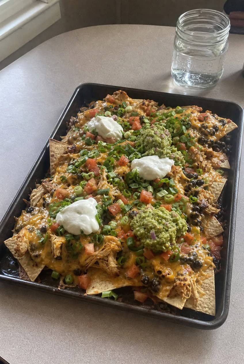 A large sheet pan loaded with golden tortilla chips topped with shredded rotisserie chicken, melted cheddar, black beans, corn, sliced jalapeños, and a swirl of sour cream, photographed on a rustic wooden table.