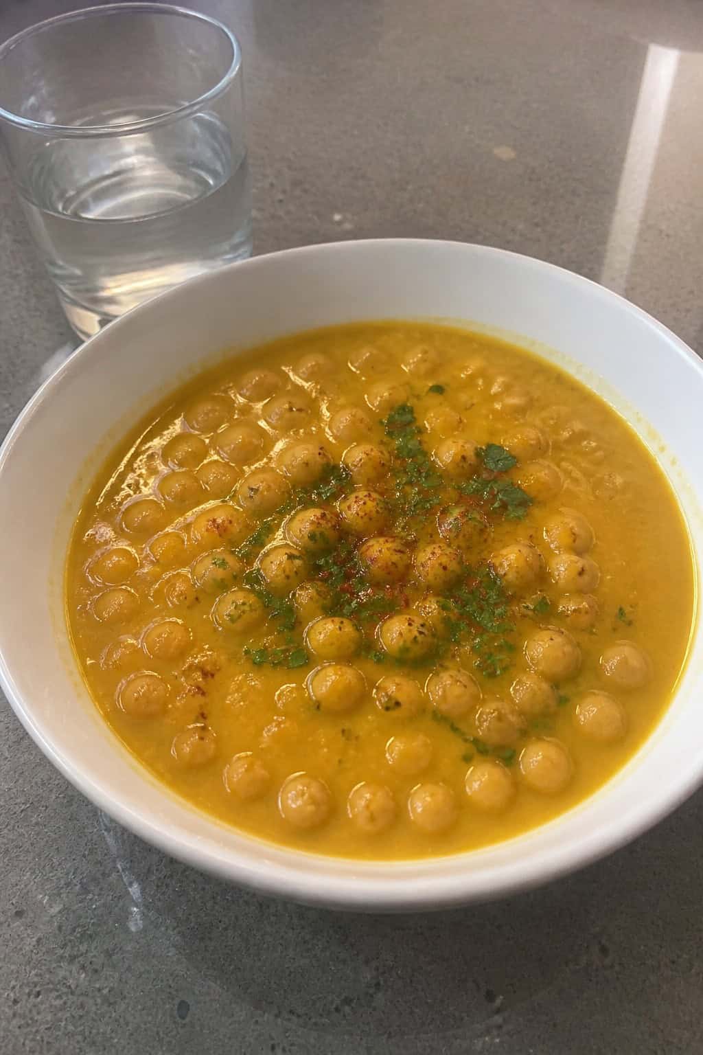 A bowl of saffron and chickpea Moroccan soup garnished with fresh herbs on a wooden table with ingredients and a spoon nearby.
