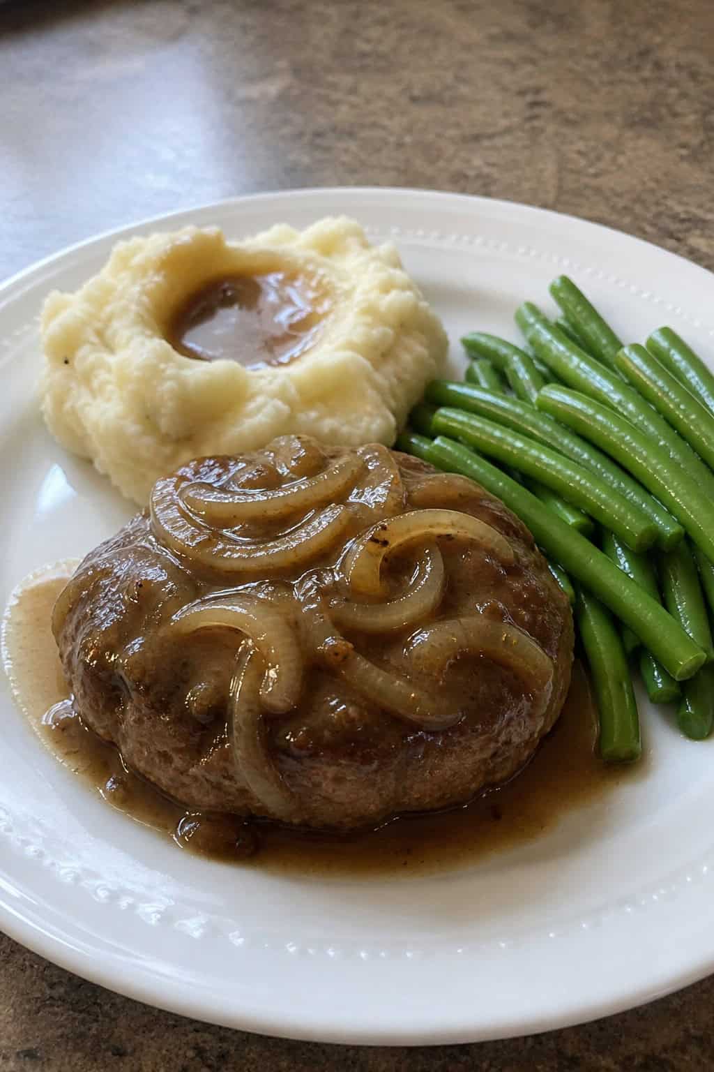 A plate of Salisbury steak with onion gravy, mashed potatoes, and green beans on a wooden table.
