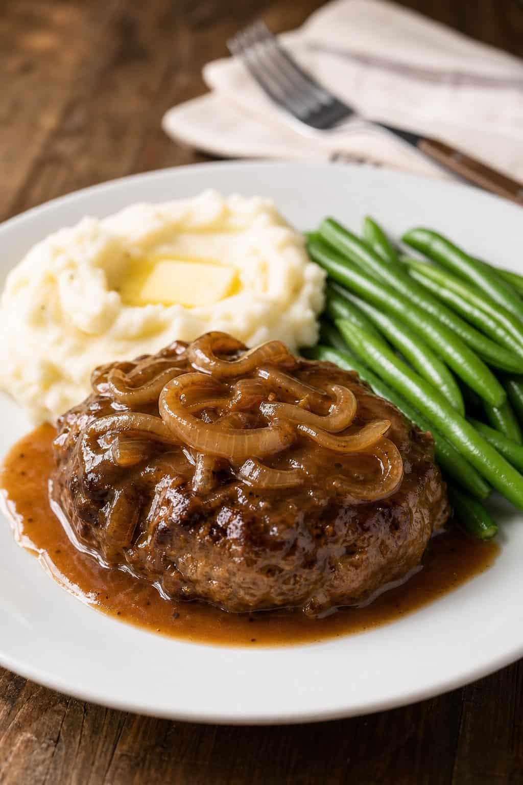 A plate of Salisbury steak with onion gravy, mashed potatoes, and green beans on a wooden table.