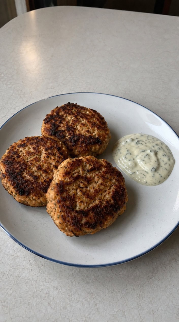 Plate of golden salmon patties with a bowl of lemon-dill sauce, garnished with fresh dill on a wooden table.