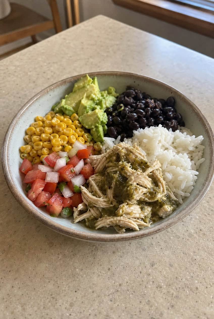 A wide bowl filled with a base of white rice topped with salsa verde chicken, black beans, roasted corn, diced avocado, shredded cheese, a dollop of sour cream, and a lime wedge, photographed on a wooden table.