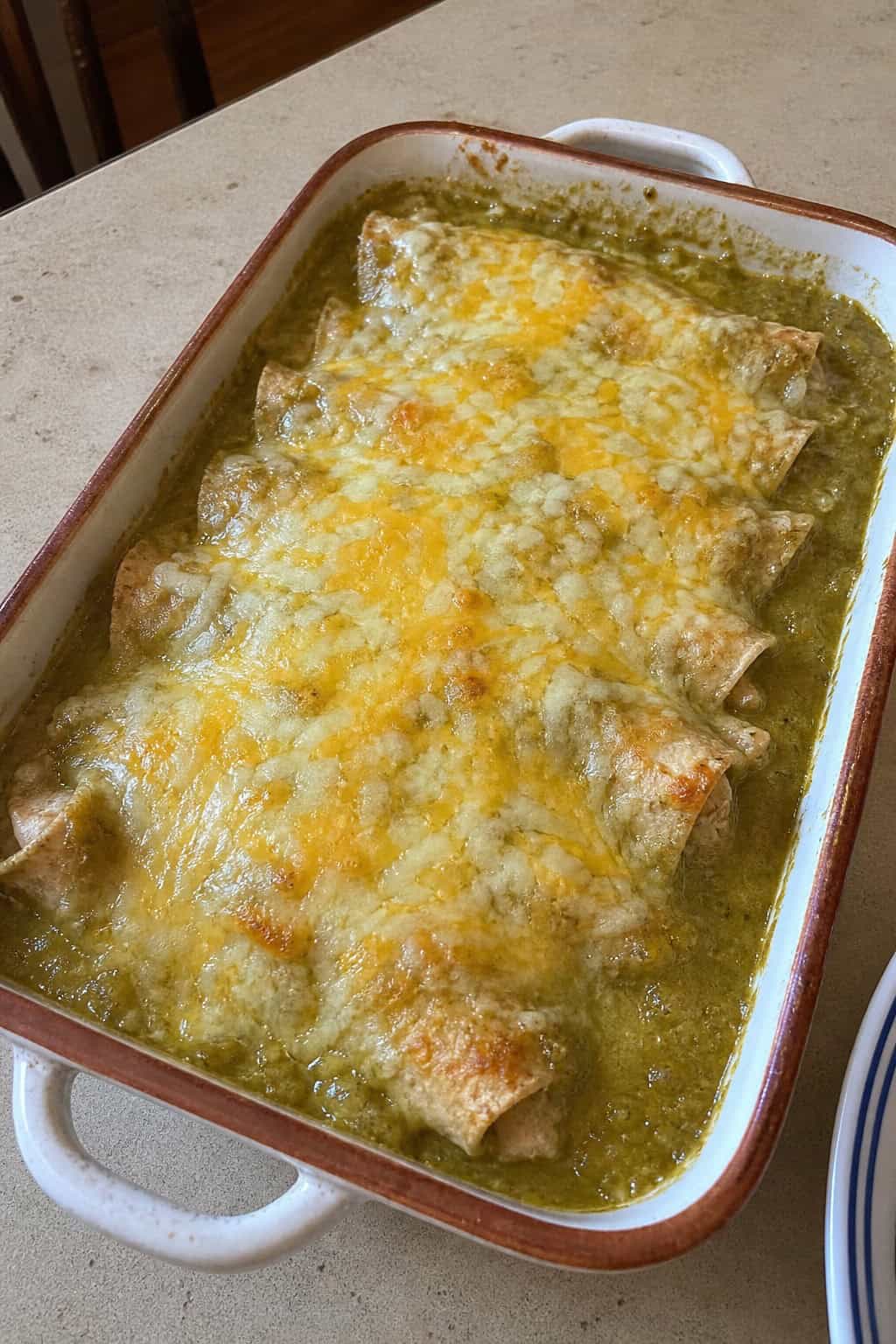 A freshly baked salsa verde chicken enchilada casserole in a white ceramic dish, garnished with fresh cilantro, with lime wedges and a small bowl of bright green salsa verde on the counter beside it.